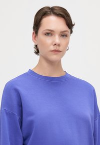 Sudadera morada hecha de tela suave, con un escote redondo, hombros caídos y un corte relajado. Textura suave, diseño minimalista.