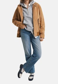 Bruine suède jas over een grijze hoodie, gecombineerd met lichtblauwe jeans en marineblauwe schoenen. Het ontwerp is casual en gelaagd.