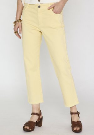 Vrouw die lichtgele cropped jeans met rafelige zomen, bruine sandalen met hak en armbanden om haar linker pols draagt tegen een effen achtergrond.