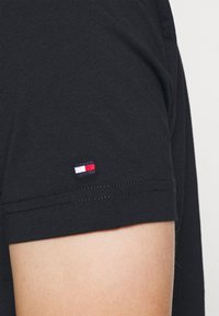 Detailní záběr na rukáv černé košile s krátkým rukávem, na kterém je poblíž lemu vyšitý malý obdélníkový logotyp v červené, bílé a námořnické modré barvě.