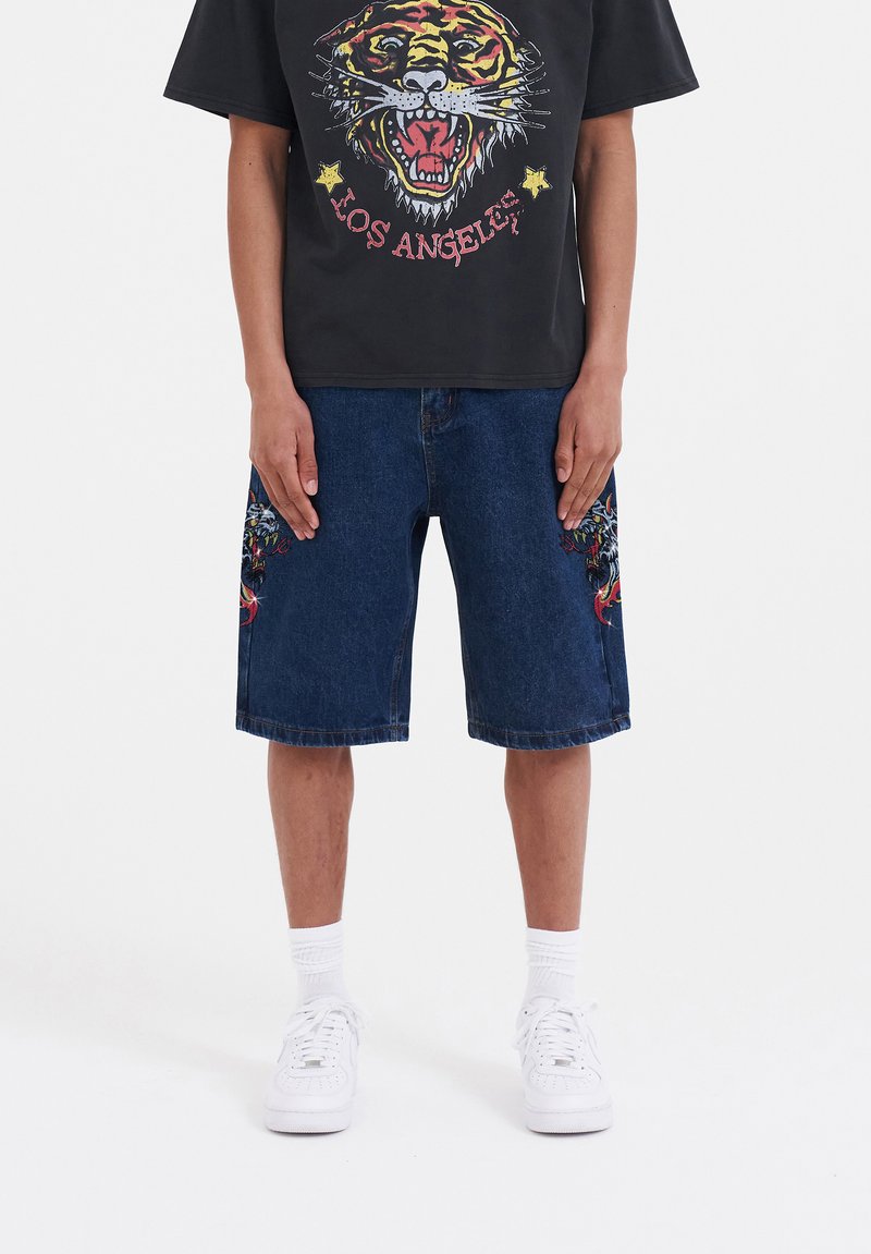 Mörkblå denimshorts med intrikata tigermönster broderade framtill och en avslappnad passform, kombinerat med en svart t-shirt med grafik och vita sneakers.