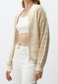 Cardigan en maille beige avec fermeture éclair et dentelle, porté sur un débardeur blanc court, associé à un pantalon blanc taille haute.