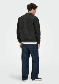 Homme portant une veste bomber noire, une chemise blanche, un jean ample bleu foncé et des baskets noires, debout de dos sur un fond uni.