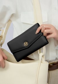 Portafoglio in pelle nera testurizzata con logo dorato, tenuto in mano accanto a una borsa in pelle crema. Il portafoglio presenta una chiusura a pattina e una forma rettangolare.