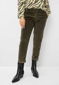 Olivgrüne Cordhosen mit Slim-Fit, umgeschlagenen Säumen, kombiniert mit schwarzen Knöchelstiefeln und einer gemusterten Bluse mit grünen und braunen Streifen.