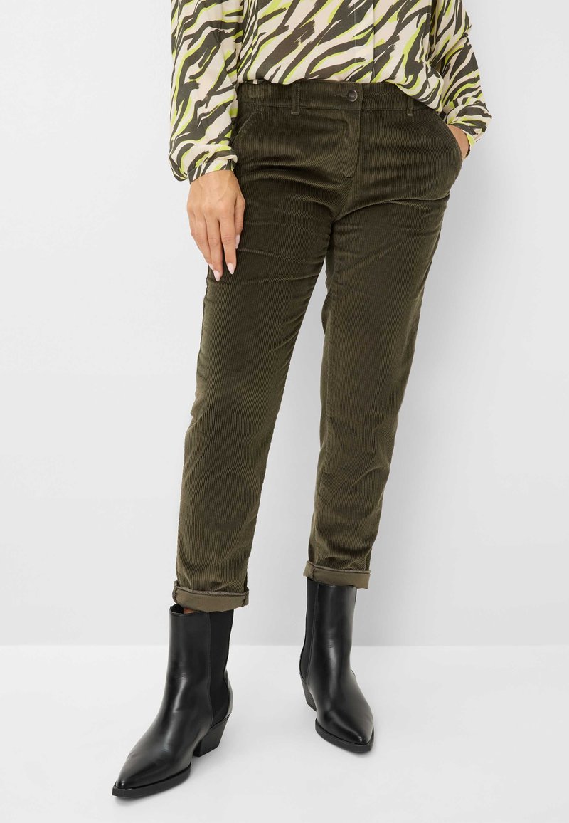 Olivgrüne Cordhosen mit Slim-Fit, umgeschlagenen Säumen, kombiniert mit schwarzen Knöchelstiefeln und einer gemusterten Bluse mit grünen und braunen Streifen.
