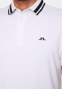 Biała koszulka polo z czarno-białym paskowanym kołnierzykiem i małym czarnym geometrycznym logo na lewej piersi.