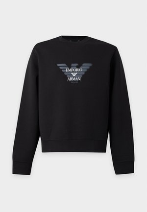 Άνδρας που φοράει μαύρο φούτερ Emporio Armani, ανοιχτόχρωμο τζιν και λευκά αθλητικά παπούτσια, στέκεται με το ένα χέρι στην τσέπη.