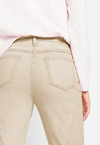 Žena oblečená v světle béžových kalhotách s zadními kapsami a v bledě růžovém topu s dlouhým rukávem, viditelná od pasu dolů a zezadu.