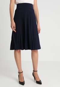 Laivastonsininen laskostettu hame, jossa on korkea joustava vyötärö. Valmistettu pehmeästä kankaasta, joka laskeutuu kauniisti polven puoliväliin asti. Yhdistetty mustiin korkokenkiin.