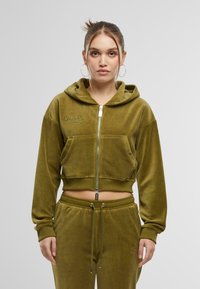 Veste à capuche cropped vert olive en velours doux, avec une fermeture éclair sur le devant, deux poches avant et un détail de logo brodé.