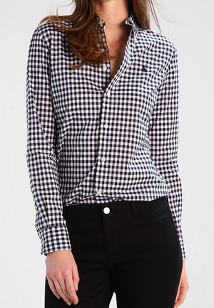 Frau trägt ein schwarz-weiß kariertes Langarmhemd, das in eine hoch taillierte schwarze Hose gesteckt ist, und steht vor einem einfarbigen Hintergrund.