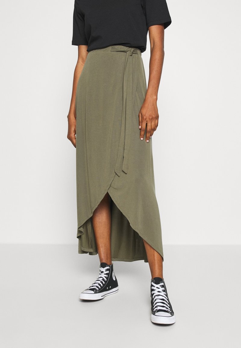 Personne portant une jupe portefeuille asymétrique vert olive avec des baskets montantes noires et un haut noir à manches courtes sur un fond uni.