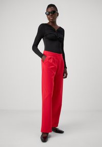 Black long-sleeve bodysuit with twist detail, paired with high-waisted, red wide-leg trousers and black flats. Accessories include black sunglasses.
