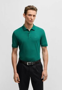 Polo shirt verde con maniche corte, caratterizzato da un colletto e un sottile logo "BOSS" sul petto, abbinato a pantaloni scuri e una cintura nera.
