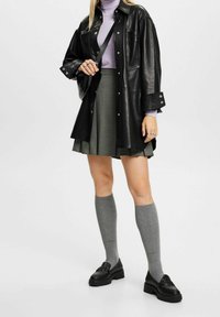 Schwarze Lederhemdjacke, lilafarbener Rollkragenpullover, grün gemusterter plissierter Rock, graue knielange Socken und schwarze Loafers mit dicken Sohlen.