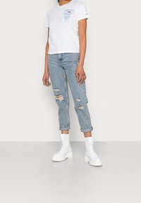 Bílá bavlněná tričko s modrým grafickým textem, světlé modré roztrhané džíny a bílé šněrovací kotníkové boty s tlustou podrážkou.