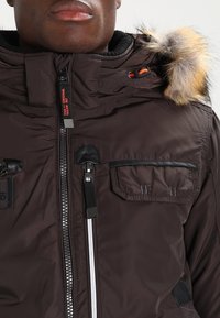 Brun pufferjakke med faux fur hætte, lynlås foran, bryst- og sidelommer samt sorte detaljer. Textureret stof med blank finish.