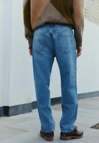Jeans in denim blu chiaro con una vestibilità comoda, caratterizzati da due tasche posteriori e una texture liscia, abbinati a calzature marroni e un maglione a fantasia.