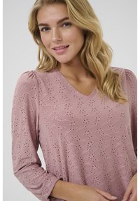 Helles rosa Blusenshirt mit V-Ausschnitt, langen Ärmeln und puffierten Schultern. Verziert mit gestickten Blumenmustern und ausgeschnittenen Akzenten. Glatte Textur.