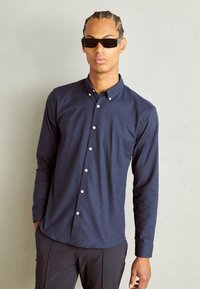 Navy long-sleeve button-up shirt made of smooth fabric, featuring a pointed collar and contrasting white buttons, worn with dark pants.
