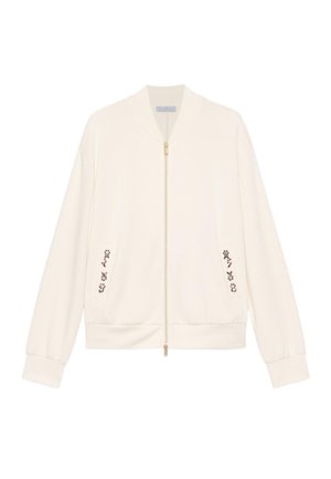 Cazadora tipo bomber de color crema con cremallera frontal y pequeño bordado floral cerca de los bolsillos, mostrada sobre un fondo blanco.