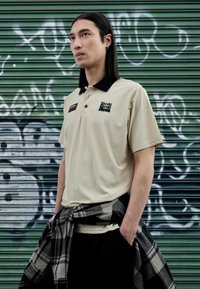 Beige polo shirt with a black collar, featuring a logo on the chest. Model is wearing black pants and a black-and-gray plaid shirt tied around the waist.