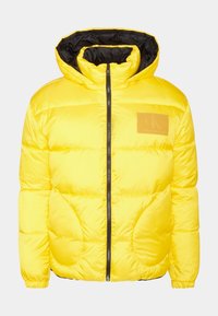 Gul puffjacka med svart inredning, dragkedja i fram och Calvin Klein-logotyp i brunt på vänster bröst.