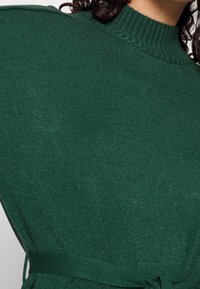 Pull vert en maille avec un col montant côtelé, des épaules structurées et un détail à la taille noué, présentant une surface texturée et une coupe ajustée.