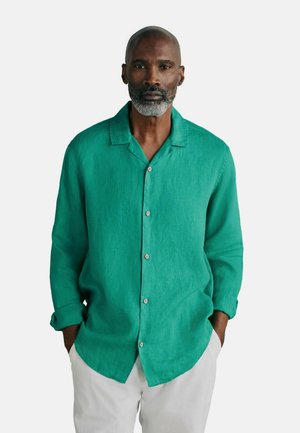 Muž so sivou bradou, oblečený v dlhom tyrkysovom košeli na gombíky a svetlých nohaviciach, stojaci s rukami vo vreckách.