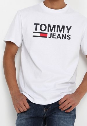 Mężczyzna ubrany w białą koszulkę z czarnym logo „TOMMY JEANS” i wzorem flagi w kolorach czerwonym, białym i niebieskim, zestawioną z niebieskimi dżinsami.