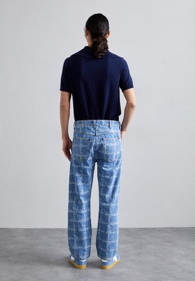Navy knitted polo shirt, light blue denim jeans with white cursive text pattern, white shoes with a beige sole, viewed from the back.