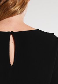 Černý oděv s kulatým výstřihem a malým průstřihem na zádech, který je zajištěn knoflíkem ze stejné látky. Hladká struktura.