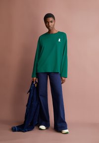 Groene lange mouwen shirt met een wit lintlogo. In combinatie met donkerblauwe flared jeans en casual witte schoenen met groene accenten.