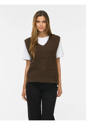 Junge Frau mit langen Haaren, die eine braune gestrickte Weste über einem weißen kurzärmligen Hemd und schwarze Hose vor einem schlichten Hintergrund trägt.