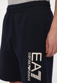 Navy cotton shorts with an elastic waistband, featuring a beige printed logo saying "EA7 EMPORIO ARMANI" on the left leg.