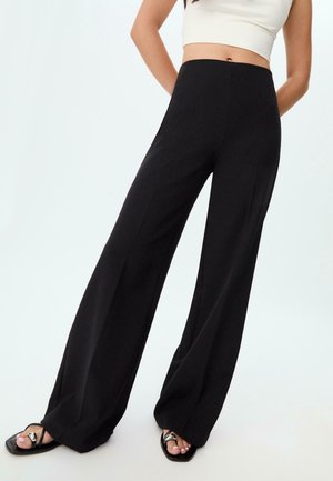 Vrouw draagt zwarte, hooggetailleerde broek met wijde pijpen, gecombineerd met zwarte sandalen met vierkante neus en een wit cropped tanktop.