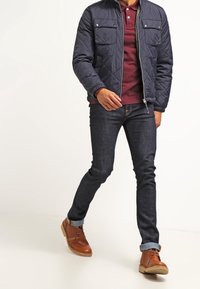 Marineblauer, gesteppter Mantel mit zwei Brusttaschen, kastanienfarbendes Poloshirt, dunkle Jeans und braune Ankle Boots mit hellen Sohlen. Lässiges Outfit.