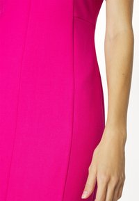 Aangemeten jurk van felroze stof, met een gladde textuur en zijzak. Close-up van de jurk met een hand op de stof.