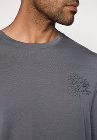 Grå kortermet skjorte med rund hals. Har svart blomsterbroderi på venstre bryst og et mykt, teksturert stoff.