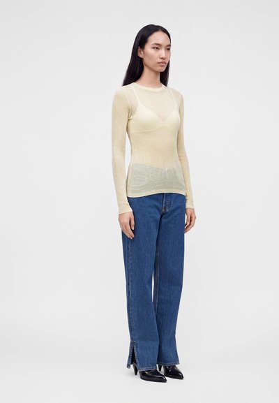 Femme aux longs cheveux foncés portant un haut beige transparent à manches longues, un jean bleu avec des fentes sur le devant et des escarpins noirs à bout pointu, debout devant un fond uni.
