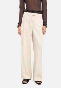 Jasnobeżowe spodnie o szerokich nogawkach z teksturowanej tkaniny, wyposażone w pasek przewleczony przez szlufki i przednie kieszenie. Noszone z czarnymi sandałami typu slide.