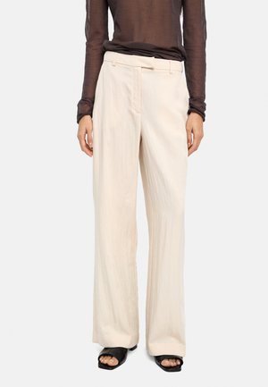 Jasnobeżowe spodnie o szerokich nogawkach z teksturowanej tkaniny, wyposażone w pasek przewleczony przez szlufki i przednie kieszenie. Noszone z czarnymi sandałami typu slide.