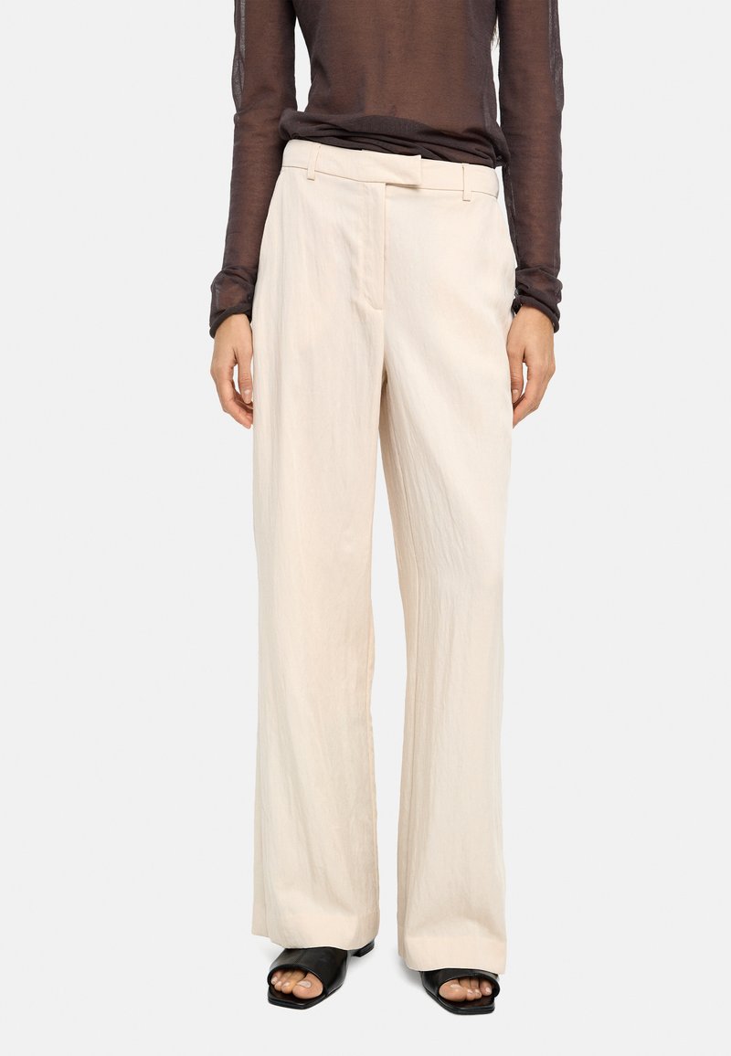 Jasnobeżowe spodnie o szerokich nogawkach z teksturowanej tkaniny, wyposażone w pasek przewleczony przez szlufki i przednie kieszenie. Noszone z czarnymi sandałami typu slide.