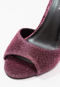 Gros plan sur une chaussure à talon haut ouverte sur les orteils, recouverte de paillettes rose foncé étincelantes avec une semelle intérieure noire.