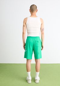 Shorts en coton verts avec une taille élastique et une poche latérale, dotés d'un détail logo blanc, associés à un débardeur blanc et des chaussettes.