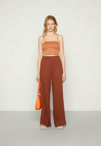 Pantalon large marron à taille haute, associé à un crop top à motifs orange. Le mannequin tient un petit sac à main orange.