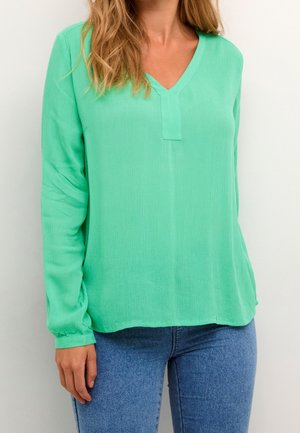 Kobieta w miętoszmaragdowej bluzce z dekoltem w serek i długim rękawem, zestawiona z niebieskimi dżinsami, na tle białej, jednolitej ściany.