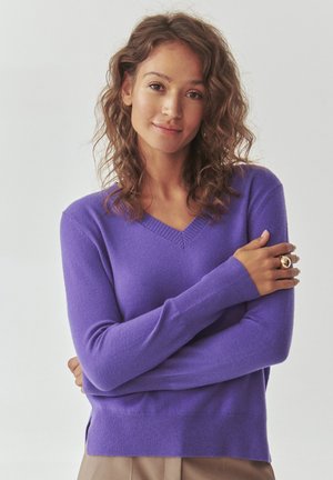 Maglione viola a V, con maniche lunghe, polsini a coste e spacchi laterali. Realizzato in materiale morbido, presenta una texture liscia.