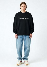 Felpa nera con testo bianco "HO PARLATO DI TE A CHATGPT", abbinata a jeans azzurri chiari e sneakers multicolori. Texture del tessuto liscia.